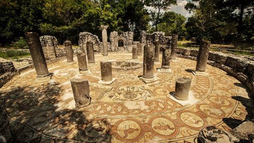 Ruins of stone columns and walls ontop of a patterned floor, surrounded by vegetation