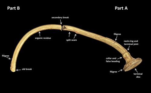 Colour photograph of torc, shining gold against a black background with all features indicated (namely old break, filigree, organic residue, secondary break, split seam, neck and ring terminal joint, collar and false beading and terminal disc)